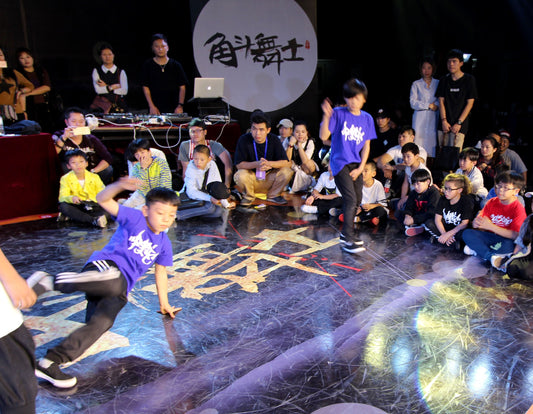 Two young b-boys in purple shirts battle in a circle at a breaking event in Fujian, China, with kids and adults watching from the floor.
