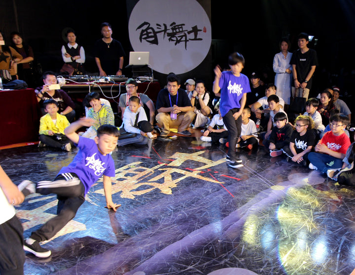 Two young b-boys in purple shirts battle in a circle at a breaking event in Fujian, China, with kids and adults watching from the floor.