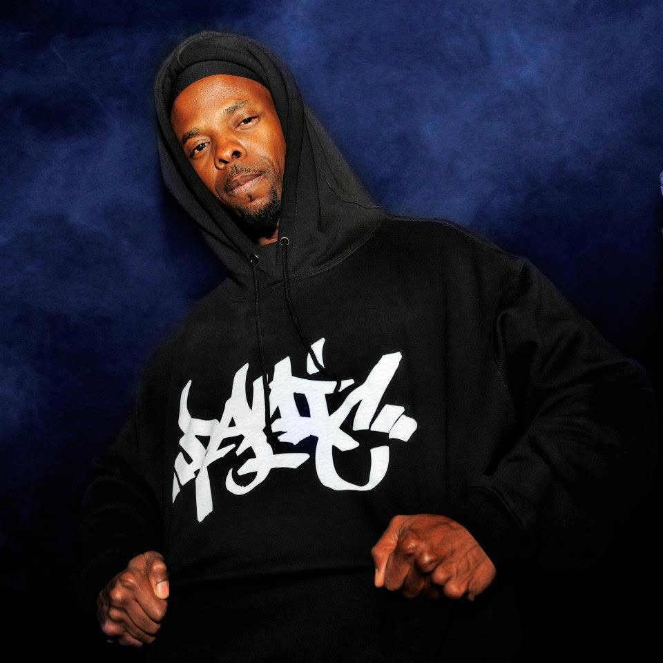 DJ Lord of Public Enemy in a black Panic 39 hoodie with the hood up and the white graffiti tag logo on the chest, pointing at the camera against a blue studio backdrop.